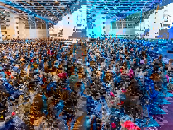 Large conference room with three giant screens along right hand wall and stage set up below the middle screen.  Audience seated in rows in front of stage. 