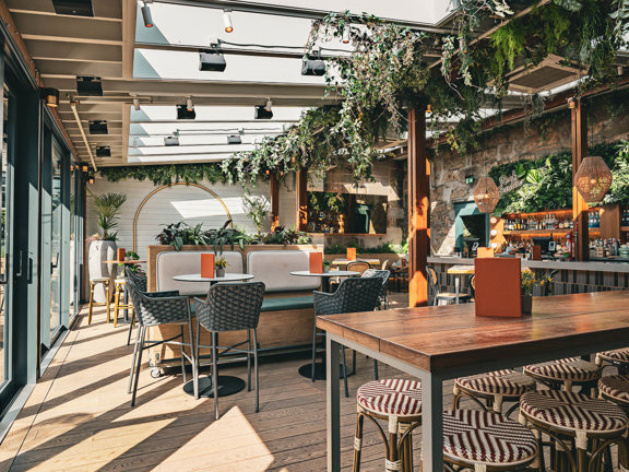Interior of Kong bar in Glasgow featuring a cosy, modern space with wooden tables and stools, hanging greenery, and a well-stocked bar against a stone wall backdrop.