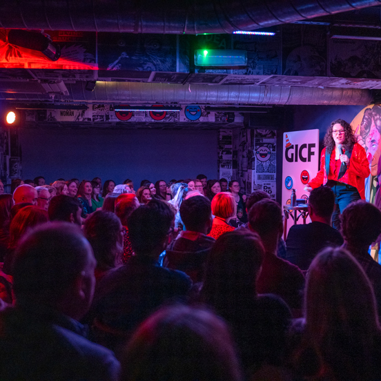 A comedian performs to a small audience, next to a Glasgow International Comedy Festival sign.