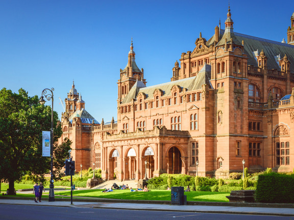 An exterior view of part of the Kelvingrove museum on a sunny day, shows the ornate, Victorian red-sandstone facade of the main entrance. The building is heavilyadorned, the main entrance is dominated by 3 large archways at the top of a few steps. The roof line is decorated with pinnacles, buttresses and turrets. In front of the museum, a sumptuous green lawn, shrubs and trees line the pavement and road in the foreground of the image. A pedestrian crossing and an ornate lamppost are also visible.