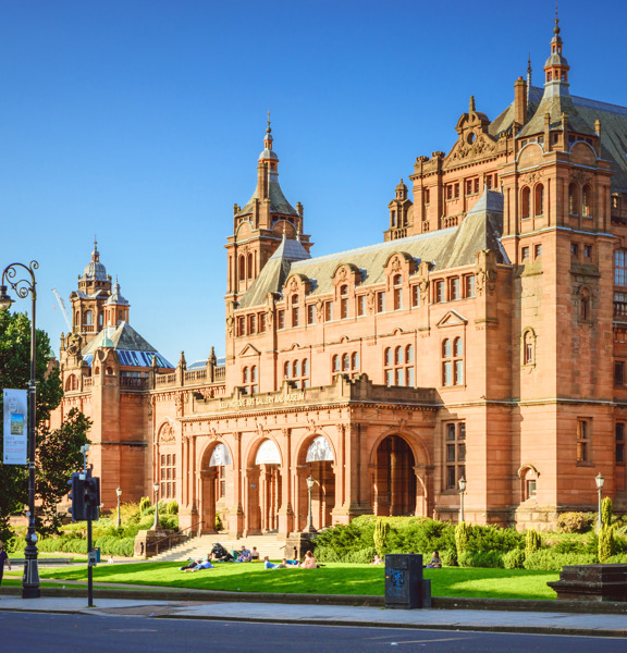 An exterior view of part of the Kelvingrove museum on a sunny day, shows the ornate, Victorian red-sandstone facade of the main entrance. The building is heavilyadorned, the main entrance is dominated by 3 large archways at the top of a few steps. The roof line is decorated with pinnacles, buttresses and turrets. In front of the museum, a sumptuous green lawn, shrubs and trees line the pavement and road in the foreground of the image. A pedestrian crossing and an ornate lamppost are also visible.