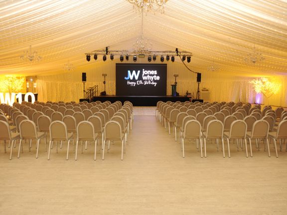 Large marque set up with rows of chairs and a stage and TV at the far end
