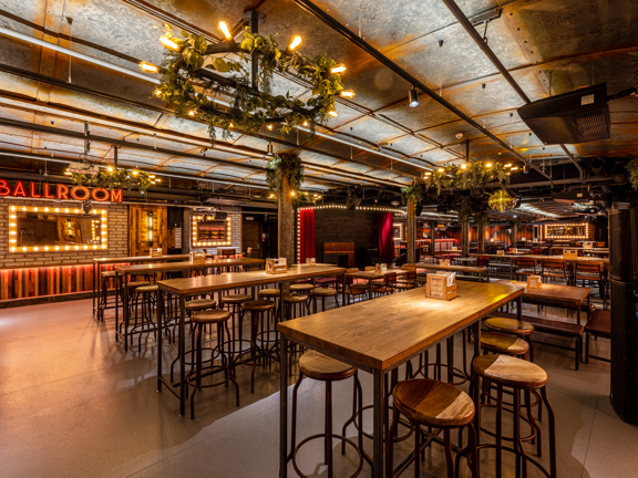 Large event room with a theatre style. Red velvet drapes and light bulbs surround the stage, ambient lighting and large mirrors with lightbulbs surrounding them. Illuminated sign reads Ballroom. 