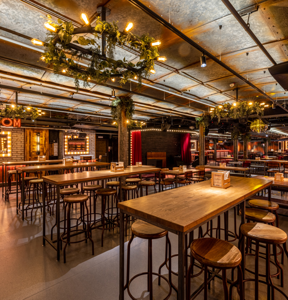 Large event room with a theatre style. Red velvet drapes and light bulbs surround the stage, ambient lighting and large mirrors with lightbulbs surrounding them. Illuminated sign reads Ballroom. 