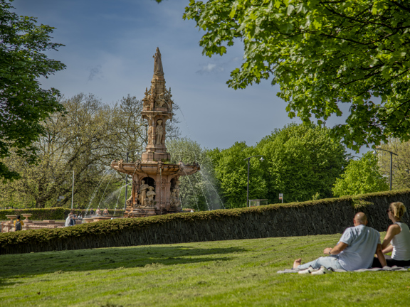 A couple lazing in the sunshine near the large terracotta, five-tier, French Renaissance-style Doulton Fountain in Glasgow Green