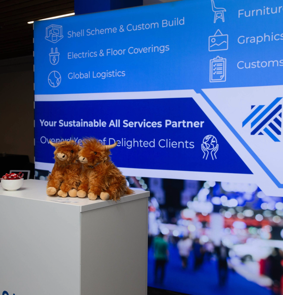 Exhibition stand with blue backdrop that reads Cameron Event Logistics, your sustainable all services partner. a table sits in front with two highland cow stuffed toys on it. 