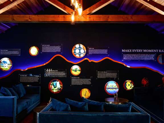 A dark and moody room with dark blue walls and dark blue velvet sofas. Circular pictures are back lit along the wall at varying heights. A sign reads Make Every Moment Raer. 