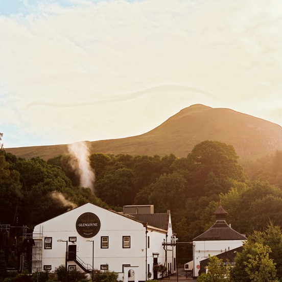 An exterior view of the Glengoyne Distillery shows a 3 storey building with white rendering and a gabled roof, dwarfed by the trees and hills behind it. There is a neighbouring smaller building obscured by trees. The large building has a large black circle on it with white lettering inside it reading "Glengoyne." The sky is bright giving a golden glow to the large trees, the striking outline of a hill and the sky. It also highlights the steam or smoke rising from the main building. 