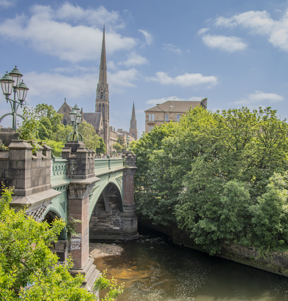 Sunny view of the green metal Kelvin Bridge surrounded by greenery and a tall church spire in the background