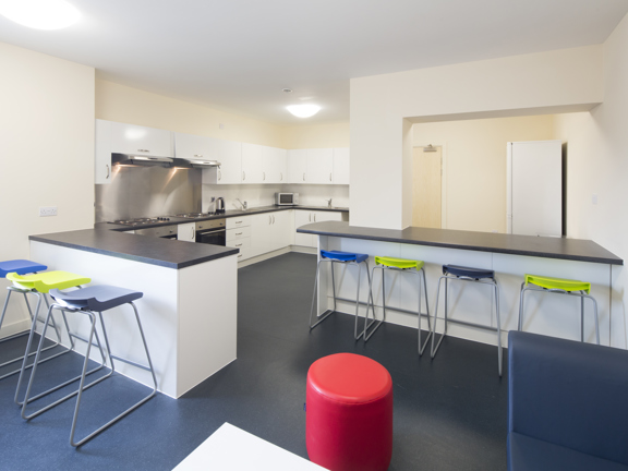 Large communal kitchen with fitted white units, two ovens and a fridge freezer. Two island units with blue, yellow and grey bar stools provide seating for dining. 