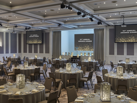 Hotel meeting room set up for an awards ceremony. Round tables set with candles, crockery and beige table linen. Three large screens are situated across the far wall and all read 10th Annual Awards Dinner. 