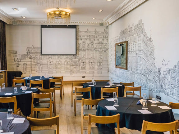 An internal view from the Corinthian Club shows a room with wooden chairs arranged around several round tables with black table cloths. The room is bright with the edge of a window visible on the left-hand side and light fittings in the ceiling. The walls are decorated with white wallpaper illustrated with a black outline of the Corinthian Club's exterior and surrounding street scene. A projection screen covers much of the far wall and speakers are also visible.