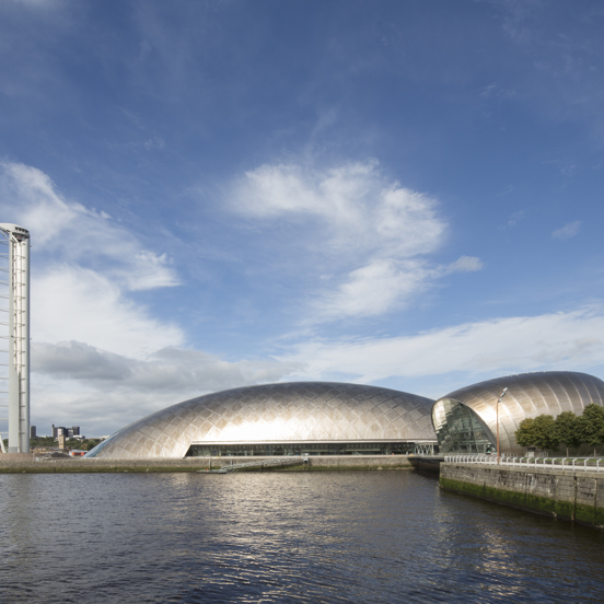 View across the River Clyde of the tall metal spire of the Glasgow Tower, of the crescent-shaped metal structure of the Glasgow Science Centre and of the round glass fronted IMAX theatre