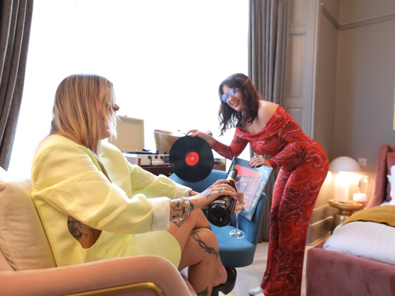 Two woman in a hotel bedroom drinking champagne, selecting music for the record player and dressed for a night out. 