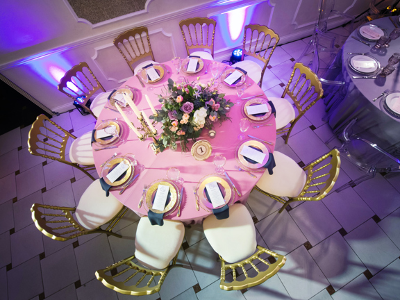 View of a 10 seater dining table set with pale pink tablecloth, flowers, candles, gold plates surrounded by cream and gold chairs. 