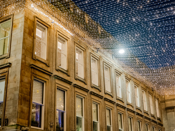 Georgian style blonde sandstone building with strings of fairly lights suspended from the roof across to buildings not in the shot. 
