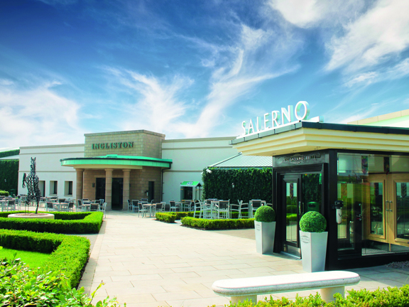 An exterior view of the Ingliston Country club shows a paved stone footpath lined with a low box hedge. The pathway leads to 2 single-storey buildings, lettering over the closest door reads, "SALERNO." At the far end of the path is a low, flat roofed building. It has sandstone facing on its entrance and 4 stone pillars holding up a semi circular awning, green-oxidised letting on the stone reads, "Ingliston." Small wooden tables and chairs surround the entrance.