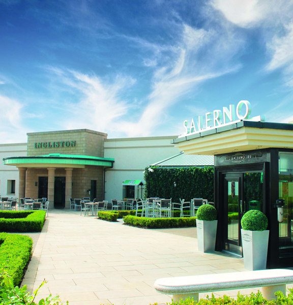 An exterior view of the Ingliston Country club shows a paved stone footpath lined with a low box hedge. The pathway leads to 2 single-storey buildings, lettering over the closest door reads, "SALERNO." At the far end of the path is a low, flat roofed building. It has sandstone facing on its entrance and 4 stone pillars holding up a semi circular awning, green-oxidised letting on the stone reads, "Ingliston." Small wooden tables and chairs surround the entrance.