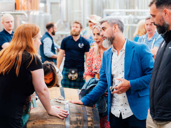 A woman stands opposite both with their hand placed on a whisky barrel. Others stand around listening to the conversation. A man in the background is wearing a kilt. 