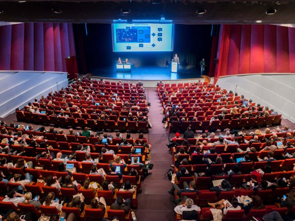 Large conference room with screen and stage at front. Audience seated in rows on red chairs.