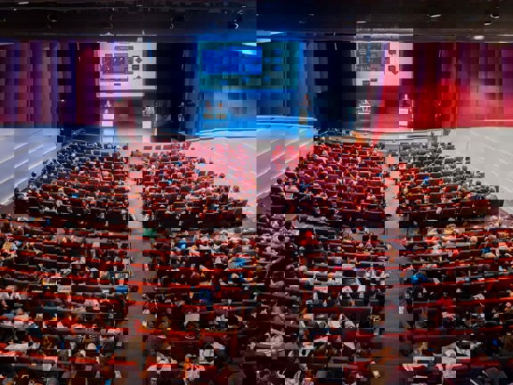 Large conference room with screen and stage at front. Audience seated in rows on red chairs.