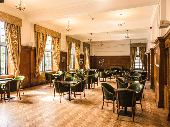 Large period room with parquet flooring, wooden panelling and five large windows along one length. There are green leather armchairs around wooden tables throughout the room.
