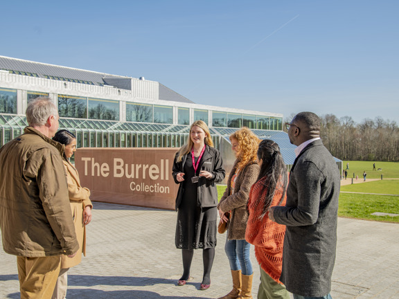 Group of people standing outside a modern red brick and glass building, a museum known at The Burrell Collection. Sun is shining, blue sky and a brick sign behind the group reads The Burrell Collection 