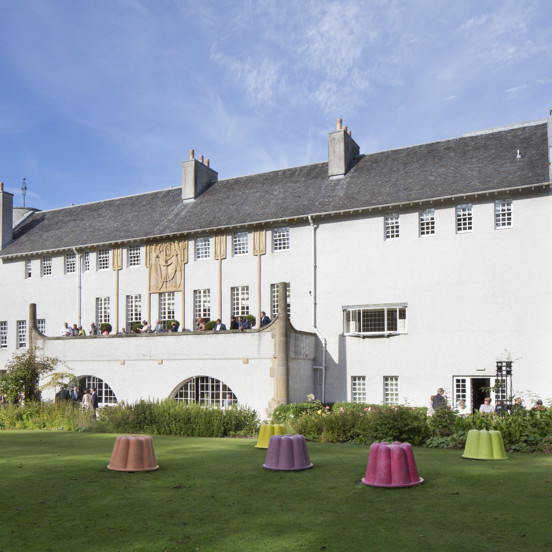 An external view of Glasgow's House for An Art Lover, a Charles Rennie Mackintosh building with 3 storeys, white rendering, blonde sandstone art-nouveau carved details and a shallow, dark peaked roof. A large balcony stretches out of the first floor into a manicured garden, there is a lawn and a wide flower bed between the viewer and the building. People sit on the terrace and mill around a french window entrance in the ground floor. Jelly mould shaped sculptures in bright colours, dot the lawn.