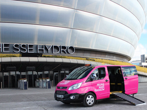 pink taxi with People Make Glasgow branding in front of a modern concert arena, with the ramp down