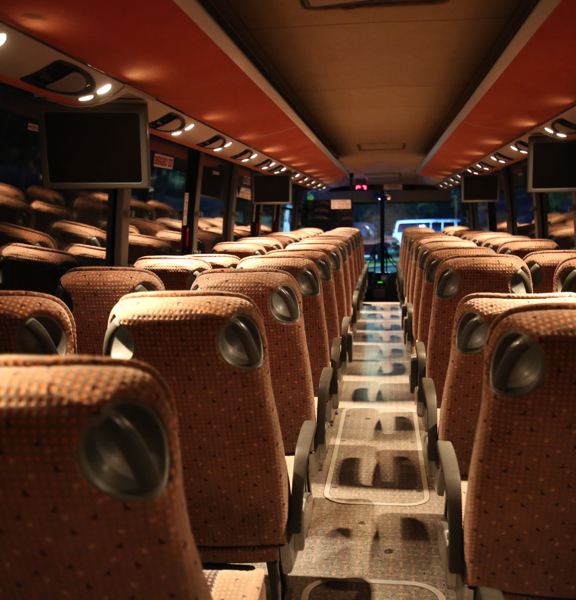 An image taken looking down the length of a luxury coach from the back seat. The seats on either side of the aisle are upholstered in orange fabric and have armrests. The floor is carpeted and the ceiling has orange panels with small spotlights above each seat. 4 small screens can also be seen attached to the ceiling. Outside is dark so the coach's interior is reflected in the large windows.