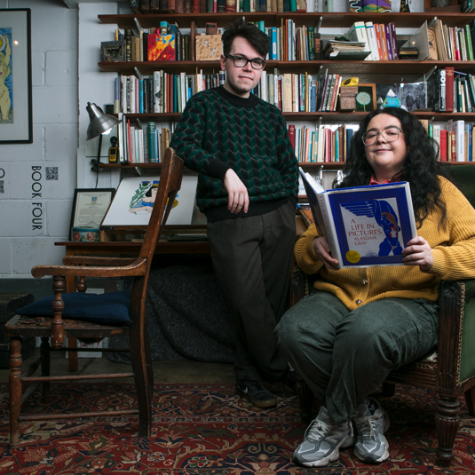 Christopher Macarthur-Boyd standing  behind  Ashley Storrie who is sitted reading a book from the archive. They are surrounded by books and drawings