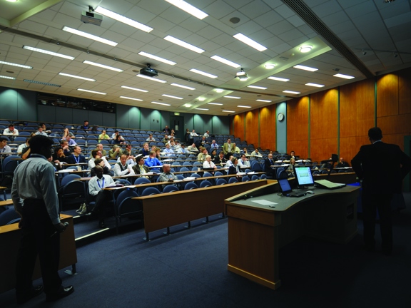 A photo of a Strathclyde University lecture theatre taken from the front of the room. The room is lit with florescent lights and the floor is carpeted, steps are clearly marked. A lecturer stands beside a wooden desk with a monitor mounted on it. Students are taking their seats in the blue-grey tiered, self writing seats, with wooden desks. 3 projectors can be seen mounted on the tiled ceiling and a clock is mounted on the far wood panelled wall. A door in the same wood can just be seen in the bottom left. 