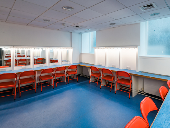 Changing room with blue floor and wrap around table with orange metal chairs, mirrors with lights above.