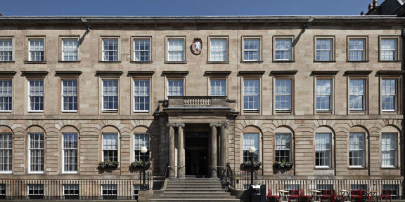 An exterior view of the Kimpton Blythswood hotel shows a grand 4-storeyed Victorian sandstone building or terrace. It is flat fronted with black railings running along its front and a grand doorway at its centre. A few steps lead up from the pavement to the doorway which is framed by Greek Ionic carved columns. The arched windows on either side are decorated with window boxes, 2 black iron, ball-topped lampposts adorn the entrance further. Small bistro tables and chairs line the pavement on the right.