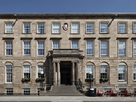 An exterior view of the Kimpton Blythswood hotel shows a grand 4-storeyed Victorian sandstone building or terrace. It is flat fronted with black railings running along its front and a grand doorway at its centre. A few steps lead up from the pavement to the doorway which is framed by Greek Ionic carved columns. The arched windows on either side are decorated with window boxes, 2 black iron, ball-topped lampposts adorn the entrance further. Small bistro tables and chairs line the pavement on the right.