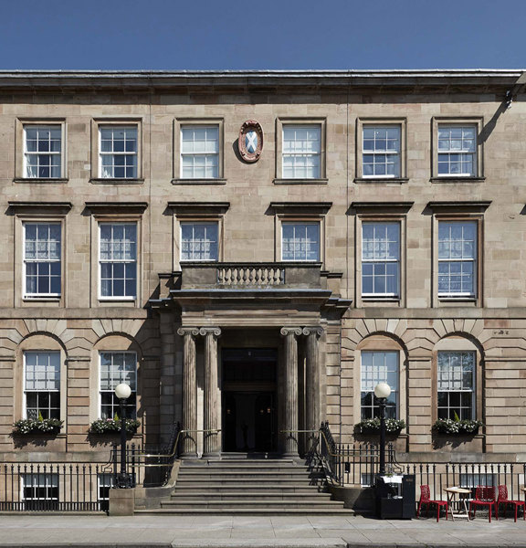 An exterior view of the Kimpton Blythswood hotel shows a grand 4-storeyed Victorian sandstone building or terrace. It is flat fronted with black railings running along its front and a grand doorway at its centre. A few steps lead up from the pavement to the doorway which is framed by Greek Ionic carved columns. The arched windows on either side are decorated with window boxes, 2 black iron, ball-topped lampposts adorn the entrance further. Small bistro tables and chairs line the pavement on the right.