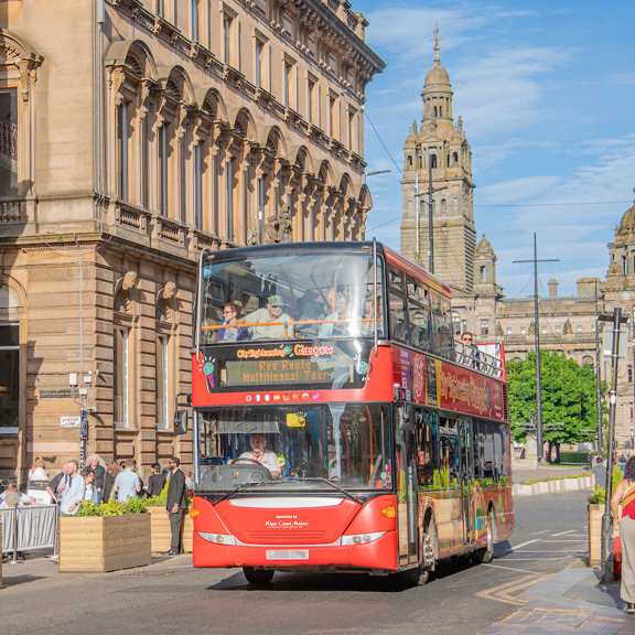 Red City Sightseeing Glasgow open-top bus travelling along St Vincent Place in central Glasgow, with outdoor café seating and the towers of the City Chambers in the background.