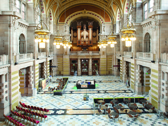 An internal view of the Kelvingrove Main Hall taken from a first storey balcony shows a cavernous, vaulted, 3 storey space adorned with carved sandstone walls, a patterned polished-stone floor and a dozen glass and golden chandaliers. A carved stone banister is visible around the entire first-storey balcony.  A huge pipe organ is visible on the balcony at the far, opposite end of the room, large wooden doors are visible beneath it. Small tables and seats are gathered together in parts of the large floor.
