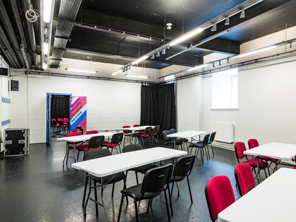Room with black floor and white walls. Several desks, each with four chairs, set up around the room.  