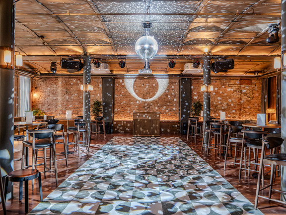 Industrial style party room with large dancefloor, glitterball and occasional table and chairs throughout. 