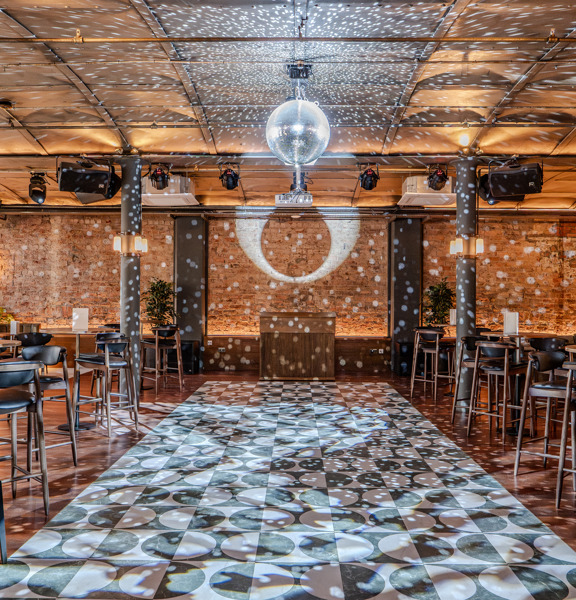 Industrial style party room with large dancefloor, glitterball and occasional table and chairs throughout. 