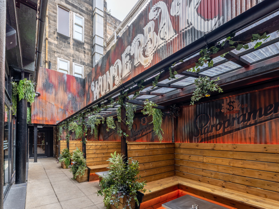 Outside terrace with corrugated iron wall with a sign reading Wunderbar, wooden bench seating and hanging greenery.