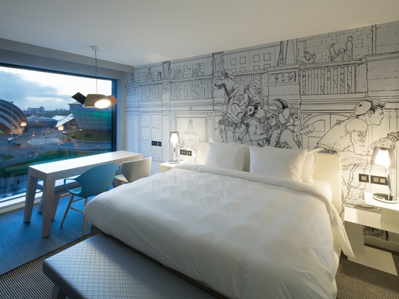 A photo of a Radisson Red bedroom, the room has a mix of wood effect flooring and black and white stripe carpet around the bed. The bed looks plush and is made with with white sheets. The headboard, contemporary bedside tables and a rectangular table in the window are white. 2 modern chairs, in blue and cream, are arranged placed side-by-side looking out of the large square window which overlooks the SEC campus. A figurative black and white line drawing takes up the right wall and ample plugs are visible. 