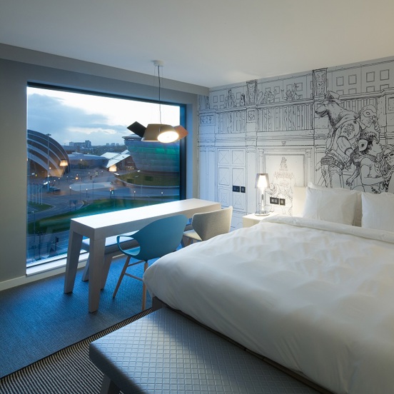 A photo of a Radisson Red bedroom, the room has a mix of wood effect flooring and black and white stripe carpet around the bed. The bed looks plush and is made with with white sheets. The headboard, contemporary bedside tables and a rectangular table in the window are white. 2 modern chairs, in blue and cream, are arranged placed side-by-side looking out of the large square window which overlooks the SEC campus. A figurative black and white line drawing takes up the right wall and ample plugs are visible. 