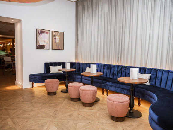 Soft seating area with a navy blue curved velvet sofa, blush pink stools and wooden tables.