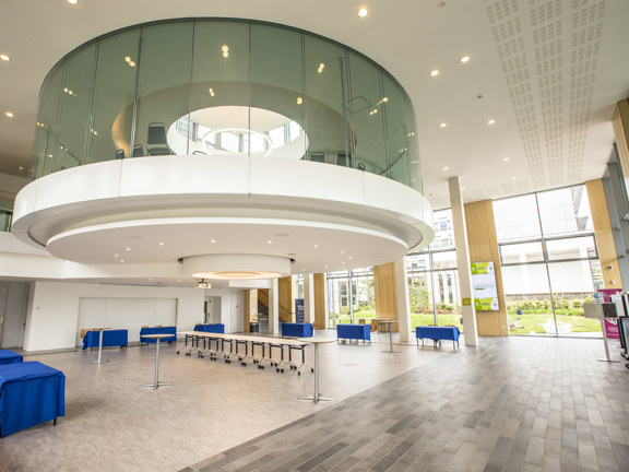 An interior view of one of Glasgow Caledonian's buildings shows a round windowed room protruding of the 2-storey ceiling of a large hall space. The large space is well lit with ceiling lights and floor to ceiling windows visible on the far external wall. In the space a variety of furniture including tables with blue cloths and chairs can be seen. Further chairs can be seen within the round glass room and on a mezzanine level behind it. Various signs, doors and a staircase are also visible in the background.