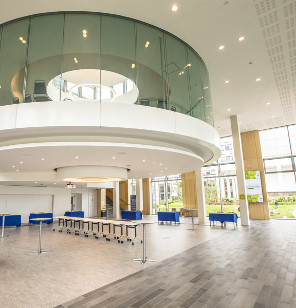 An interior view of one of Glasgow Caledonian's buildings shows a round windowed room protruding of the 2-storey ceiling of a large hall space. The large space is well lit with ceiling lights and floor to ceiling windows visible on the far external wall. In the space a variety of furniture including tables with blue cloths and chairs can be seen. Further chairs can be seen within the round glass room and on a mezzanine level behind it. Various signs, doors and a staircase are also visible in the background.