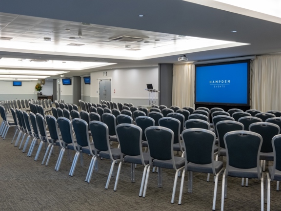 The interior of a large meeting room at Hampden Stadium. The room is decorated in muted tones and white walls, no windows can be seen on the visible walls; ceiling lights are lighting the room. Chairs are positioned into 5 curving rows facing a large projection screen and a transparent lectern with a laptop.  There are cream curtains pulled back on either side of the projection. 2 projectors can be seen on the ceiling and 3 wall mounted TV screens can be seen in the room. Multiple doorways are visible.
