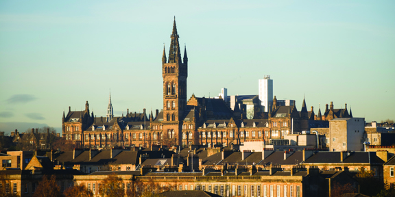 A photo of the University of Glasgow’s famous, gothic, Gilbert Scott building, taken from a distance. A wide sandstone institution with a featureful roofline including a tall, central tower topped by a latticework steeple. The sun is bright and low and reflects off some of the many lancet windows. Between the photographer and the Gilbert Scott building, the upper floors and rooves of rows of sandstone townhouses and tenements are visible, punctuated with autumnal trees and an occasional modern building.