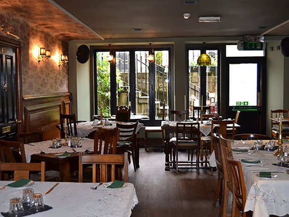 An interior image of The Butterly and the Pig's dining room. A room decorated with wooden floors and wooden panelling on the lower half of the walls. The floral wallpaper, brass sconces and embroidered white table cloths all create a "vintage" atmosphere. The far wall is lined with long, narrow, dark-framed windows; the view out of them reveals the room is on a basement level looking out at a stone staircase and black railings. There are 5 arrangements of mismatching wooden tables and chairs.